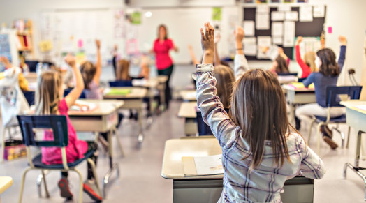 children sitting at desks in a classroom with their hands raised